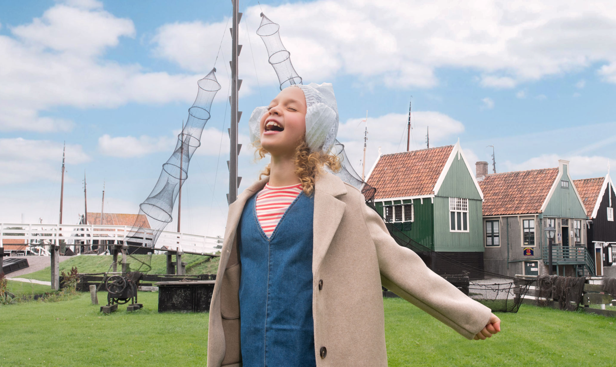 Een lachend meisje in klederdracht staat op het gras tussen oude vissershuisjes en netten